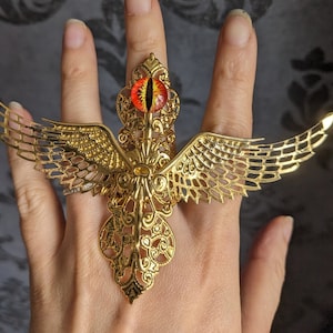 Puede incluir: Un anillo de oro con un gran diseño adornado que presenta un ojo rojo y alas. El anillo se lleva en un dedo.
