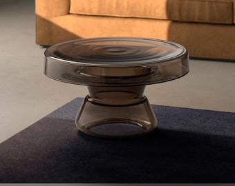 May include: A dark brown glass side table with a round top and a wide, flared base. The table is sitting on a dark blue rug in front of a brown sofa.