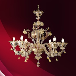 Op de afbeelding: Een gouden en heldere glazen kroonluchter met zes lichten en een bloemmotief. De kroonluchter heeft een rood en gouden middelpunt en hangt aan een ketting.