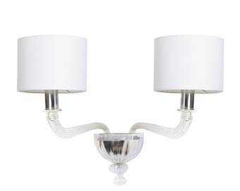 Può includere: Un'applique da parete in vetro bianco con due paralumi in tessuto bianco. L'applique ha un design curvo con un pezzo centrale di cristallo.