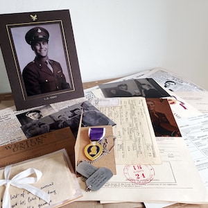Può includere: Una collezione di cimeli militari d'epoca, tra cui un ritratto incorniciato di un soldato in uniforme, una medaglia Purple Heart, targhette identificative e vari documenti e fotografie. Gli oggetti sono disposti su una superficie di legno.