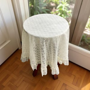 May include: A small, round wooden stool covered with a cream-colored, lace tablecloth. The tablecloth has a delicate floral pattern and a ruffled edge. The stool is positioned in front of a window, with natural light illuminating the scene.