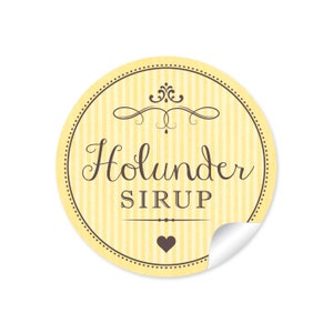 Könnte beinhalten: Ein runder gelber und weiß gestreifter Aufkleber mit einem braunen Wirbelmuster und dem Text "Holunder SIRUP" in brauner Kursivschrift. Ein kleines braunes Herz befindet sich am unteren Rand des Aufklebers.