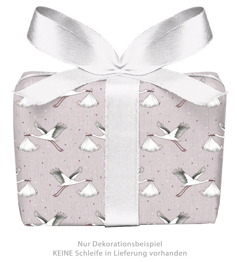 K&ouml;nnte beinhalten: Eine rosa Geschenkbox mit einem wei&szlig;en Band und einem Storchenmuster. Die Box ist mit wei&szlig;en St&ouml;rchen verziert, die B&uuml;ndel tragen, und kleinen wei&szlig;en Punkten. Der Text auf der Box lautet "Nur Dekorationsbeispiel KEINE Schleife in Lieferung vorhanden".