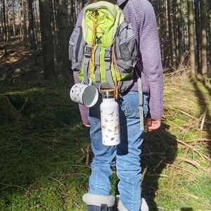 May include: A person wearing a purple sweater and a green and gray backpack with a water bottle and a mug attached to the straps. The person is standing in a forest with trees in the background.