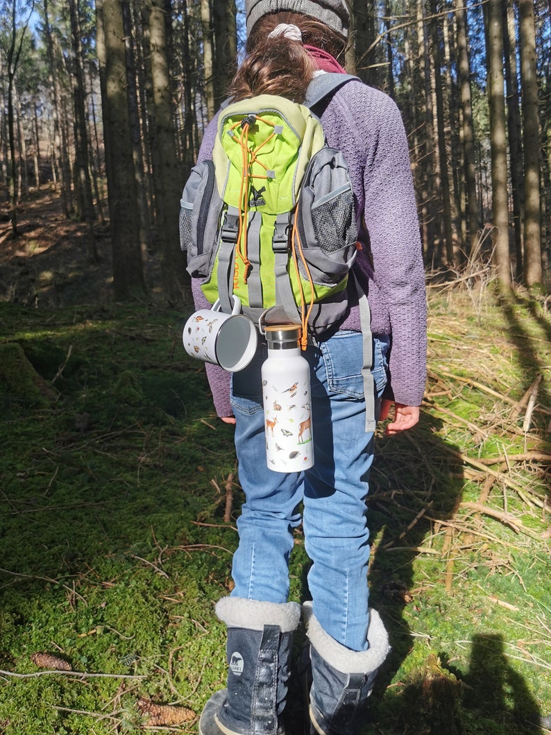 May include: A person wearing a purple sweater and a green and gray backpack with a water bottle and a mug attached to the straps. The person is standing in a forest with trees in the background.