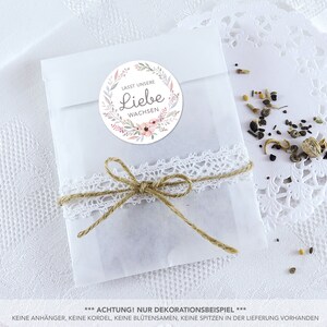 May include: A white paper bag with a floral wreath sticker that says "Lasst unsere Liebe wachsen" (Let our love grow). The bag is tied with twine and lace and has a small amount of seeds inside.