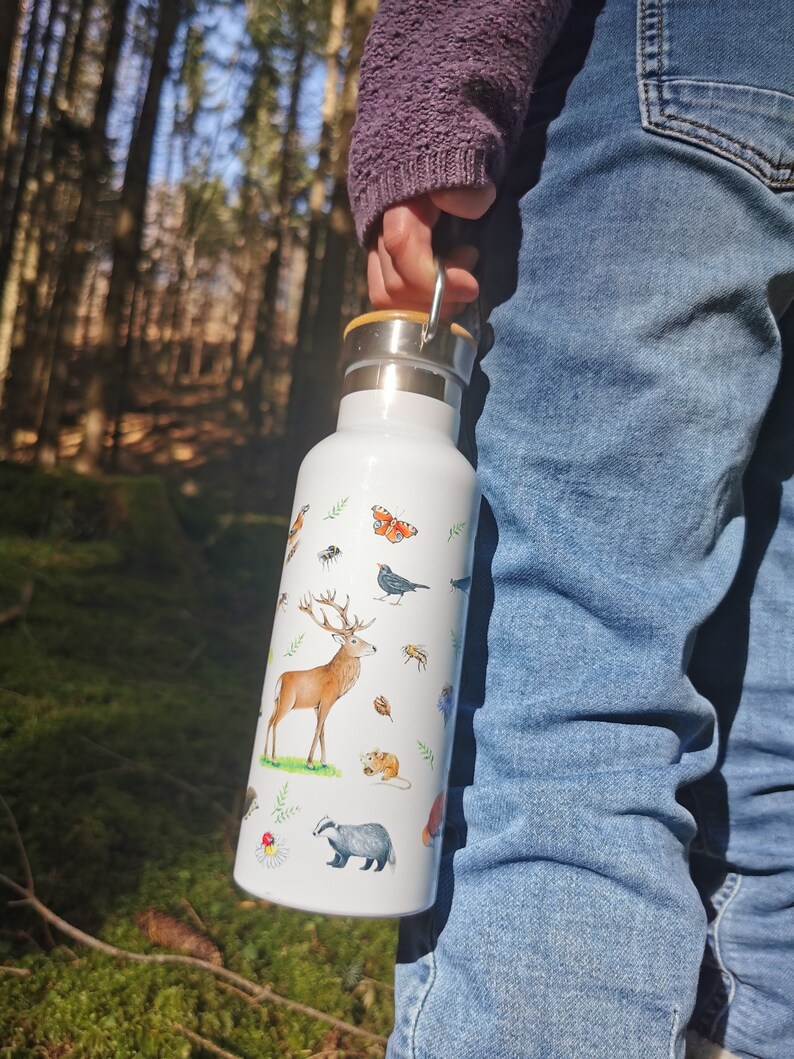 May include: White stainless steel water bottle with a wooden lid and a carrying handle. The bottle is decorated with a colorful illustration of woodland animals, including a deer, a badger, a squirrel, and a butterfly.