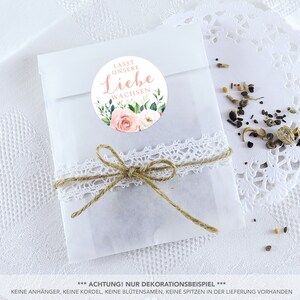 May include: A white paper bag with a pink and green floral sticker that says "Lasst unsere Liebe wachsen" (Let our love grow). The bag is tied with twine and lace.