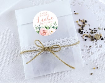 May include: A white paper bag with a pink and green floral sticker that says "Lasst unsere Liebe wachsen" (Let our love grow). The bag is tied with twine and lace.