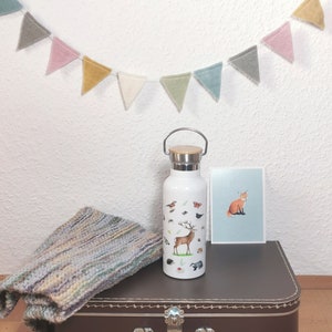 May include: White insulated water bottle with a woodland animal design. The bottle has a brown wooden lid and a brown handle. The bottle is sitting on a brown suitcase with a vintage design.