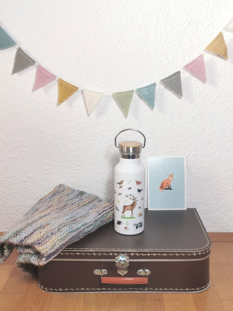 May include: White insulated water bottle with a woodland animal design. The bottle has a brown wooden lid and a brown handle. The bottle is sitting on a brown suitcase with a vintage design.
