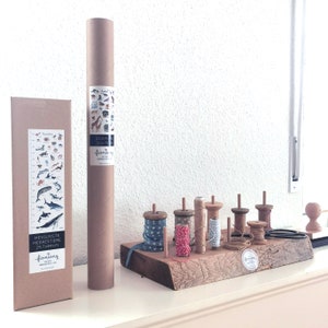 May include: Two brown cardboard tubes with illustrations of animals and the text "Zootie 50x70" and "fioniony" on them. A wooden shelf with eight spools of twine in various colors and patterns. The shelf is made of rough-hewn wood and has a natural finish.