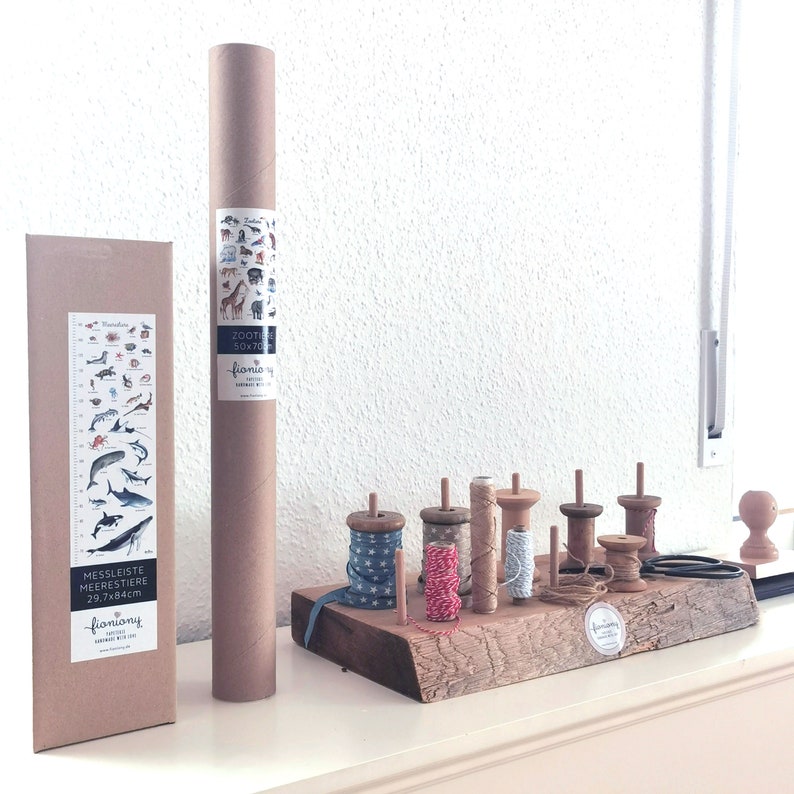 May include: Two brown cardboard tubes with illustrations of animals and the text "Zootie 50x70" and "fioniony" on them. A wooden shelf with eight spools of twine in various colors and patterns. The shelf is made of rough-hewn wood and has a natural finish.