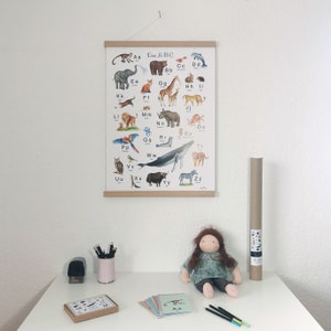 May include: A colorful alphabet poster with animals, featuring the text "Das ABC". The poster is hanging on a wall above a table with a doll, pencils, and a brown tube.