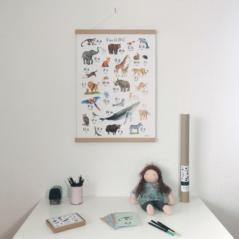 May include: A colorful alphabet poster with animals, featuring the text "Das ABC". The poster is hanging on a wall above a table with a doll, pencils, and a brown tube.