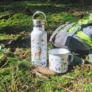 May include: A white enamel mug and a white stainless steel water bottle with a wooden lid. Both items feature a nature-themed design with illustrations of animals, insects, and plants. The items are sitting on a bed of green moss in a forest setting.