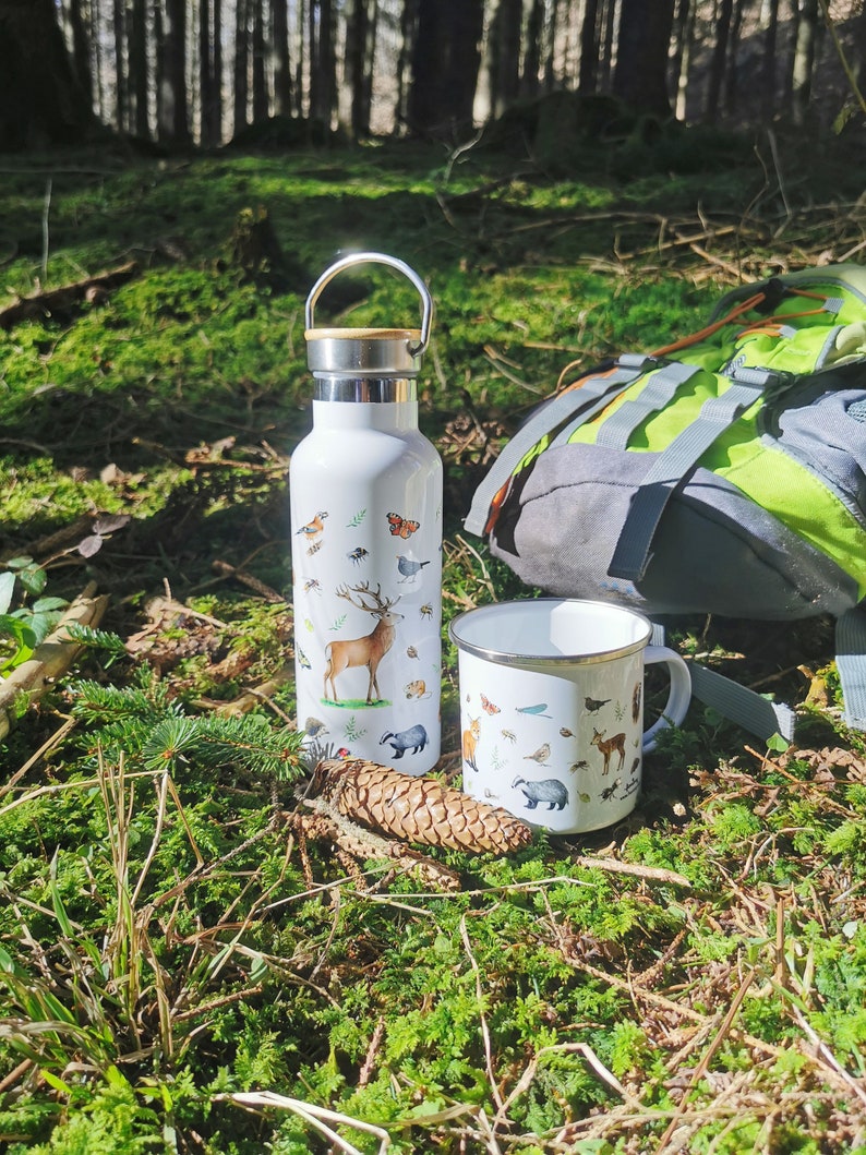 May include: A white enamel mug and a white stainless steel water bottle with a wooden lid. Both items feature a nature-themed design with illustrations of animals, insects, and plants. The items are sitting on a bed of green moss in a forest setting.