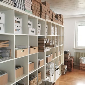 May include: A white shelf unit with multiple shelves filled with cardboard boxes, tubes, and other packaging materials. The shelves are organized and neatly stacked.