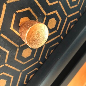 Puede incluir: Un pomo de caj&oacute;n de color dorado con una superficie texturizada sobre un fondo negro con un patr&oacute;n geom&eacute;trico de hex&aacute;gonos dorados.