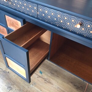 Puede incluir: Una c&oacute;moda vintage con un acabado de pintura azul oscuro y un patr&oacute;n geom&eacute;trico dorado en los cajones. La c&oacute;moda tiene un interior de madera y un caj&oacute;n est&aacute; abierto, revelando la veta de la madera.