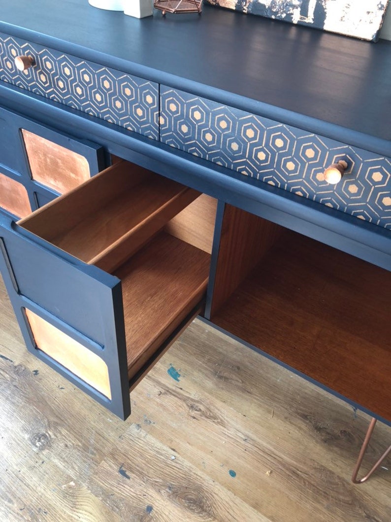 Puede incluir: Una c&oacute;moda vintage con un acabado de pintura azul oscuro y un patr&oacute;n geom&eacute;trico dorado en los cajones. La c&oacute;moda tiene un interior de madera y un caj&oacute;n est&aacute; abierto, revelando la veta de la madera.