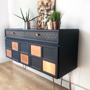 Puede incluir: Un aparador negro con detalles en cobre y cajones con estampado geom&eacute;trico. El aparador tiene cuatro puertas con paneles de cobre y se apoya en patas de horquilla.