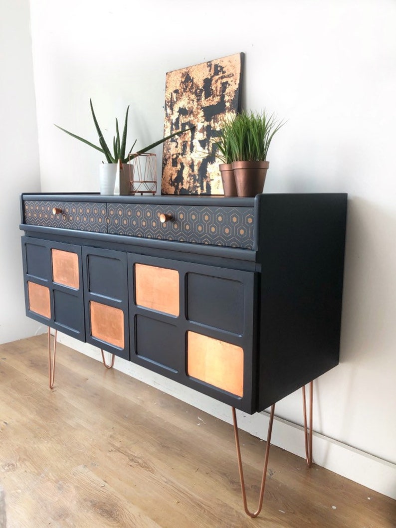 Puede incluir: Un aparador negro con detalles en cobre y cajones con estampado geom&eacute;trico. El aparador tiene cuatro puertas con paneles de cobre y se apoya en patas de horquilla.