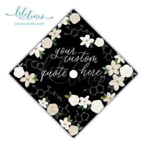 Op de afbeelding: Zwarte afstudeerhoed met witte bloemaccenten en de tekst "your custom quote here". De hoed heeft een wit gat voor de kwast en een rand van witte bloemen en groene bladeren.
