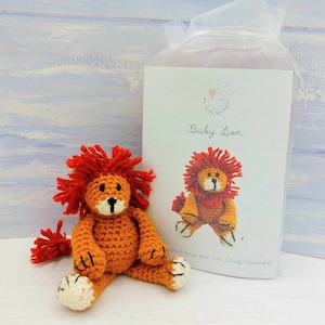 Puede incluir: Un juguete de león bebé de crochet con pelaje naranja, melena roja y patas blancas. El juguete está sentado sobre una superficie de madera azul claro junto a una tarjeta blanca con el texto "Baby Lion" y una pequeña ilustración de un león.