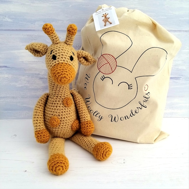 A yellow and gold crochet toy giraffe sat next to a Wee Woolly Wonderfuls crochet kit bag