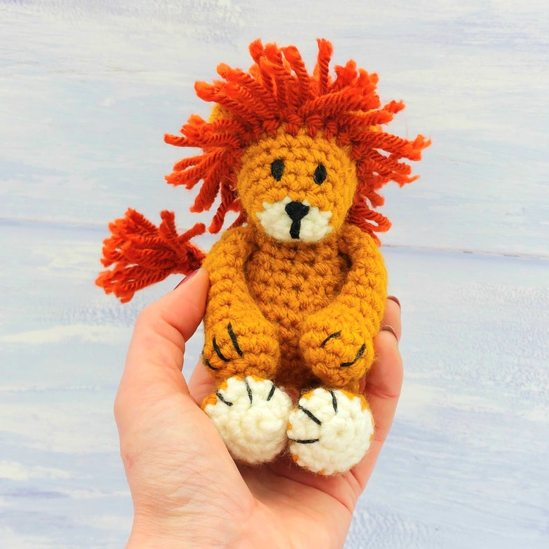 Puede incluir: Un juguete de le&oacute;n de crochet con una melena naranja brillante, un cuerpo marr&oacute;n y patas blancas con garras negras. El le&oacute;n est&aacute; sentado en una mano.