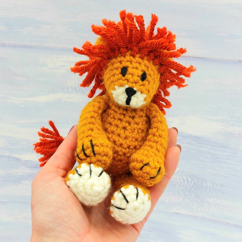 Puede incluir: Un juguete de le&oacute;n de crochet con melena naranja y cara blanca. El le&oacute;n est&aacute; sentado en una mano con las patas extendidas.