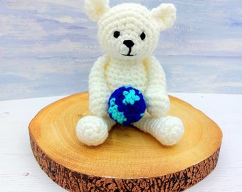 Può includere: Un orsacchiotto di peluche bianco all'uncinetto che tiene una palla di crochet blu e bianca. L'orsacchiotto è seduto su una fetta di legno.