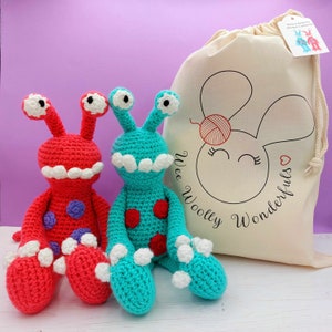 May include: Two crocheted amigurumi monster toys, one red and one teal, with white accents. The toys are sitting on a white surface in front of a white canvas bag with the text "Wee Woolly Wonderfuls" printed on it.