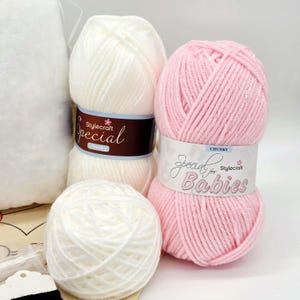 May include: Two skeins of white yarn and one skein of pink yarn. The white yarn features a dark brown label reading Stylecraft Special chunky. The pink yarn features a white label reading Special for Babies Stylecraft CHUNKY.