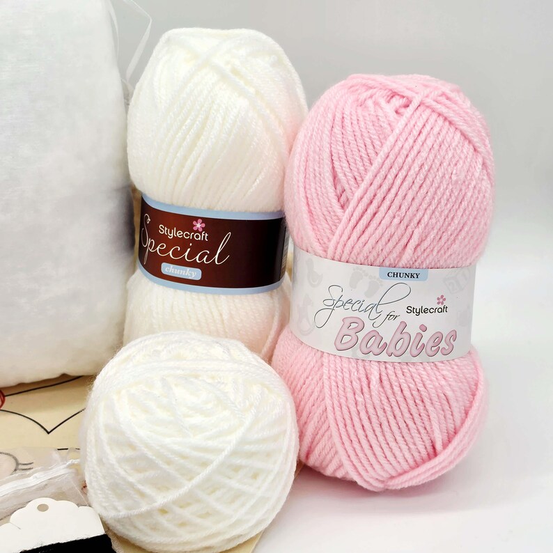 May include: Two skeins of white yarn and one skein of pink yarn. The white yarn features a dark brown label reading Stylecraft Special chunky. The pink yarn features a white label reading Special for Babies Stylecraft CHUNKY.