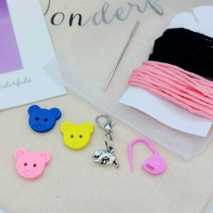 May include: A craft kit containing three bear-shaped buttons in blue, yellow, and pink, a silver-toned bear charm with a lobster clasp, a metal sewing needle, and two spools of thread in black and pink, all arranged on a light fabric surface.