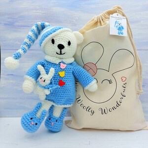 May include: A white and blue crocheted bear wearing a matching nightcap and blue pajamas with three small bear-shaped buttons. The bear holds a small white rabbit and wears blue slippers. Beside it is a beige drawstring bag labeled Woolly Wonderfuls.