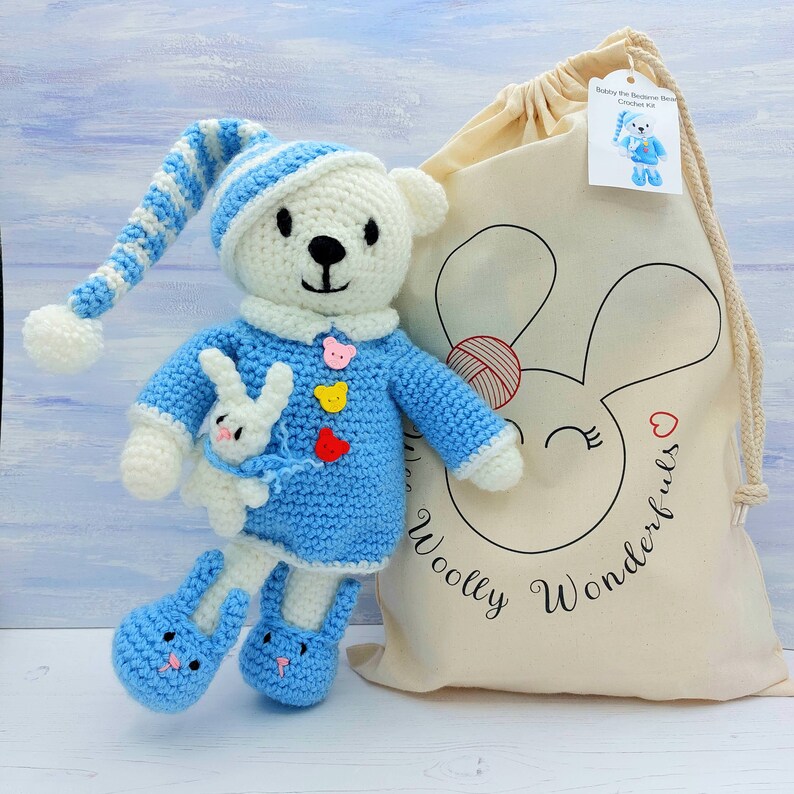May include: A white and blue crocheted bear wearing a matching nightcap and blue pajamas with three small bear-shaped buttons. The bear holds a small white rabbit and wears blue slippers. Beside it is a beige drawstring bag labeled Woolly Wonderfuls.