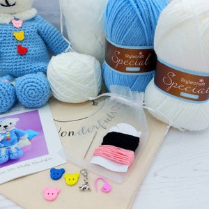 May include: A crochet kit featuring a blue bear, two skeins of Stylecraft Special chunky yarn in blue and white, a small white yarn ball, a sewing needle, black and pink embroidery thread, bear-shaped buttons, and a metal charm.