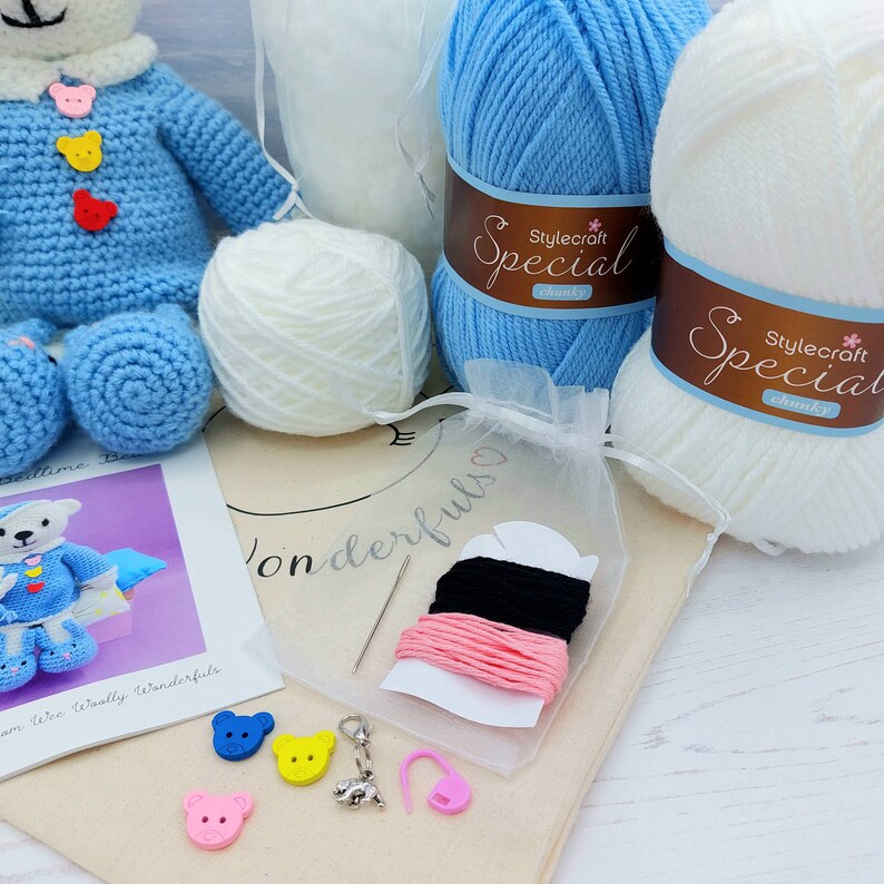 May include: A crochet kit featuring a blue bear, two skeins of Stylecraft Special chunky yarn in blue and white, a small white yarn ball, a sewing needle, black and pink embroidery thread, bear-shaped buttons, and a metal charm.