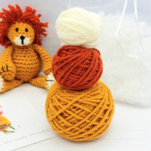 Puede incluir: Un peluche de le&oacute;n de crochet con una pila de tres ovillos de lana en naranja, amarillo y blanco. La lana es para tejer crochet.