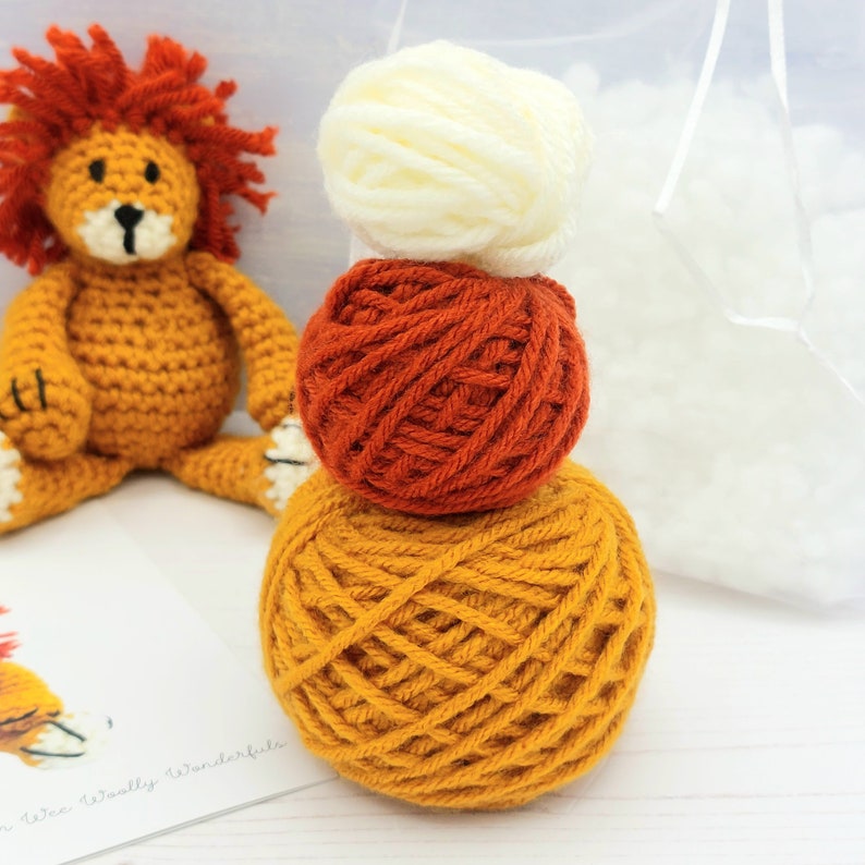 Puede incluir: Un peluche de le&oacute;n de crochet con una pila de tres ovillos de lana en naranja, amarillo y blanco. La lana es para tejer crochet.