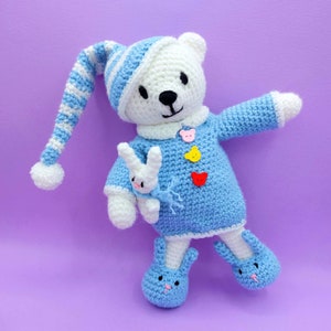 May include: A white crocheted bear wearing a blue and white striped nightcap with a pom-pom, a blue sweater with three bear-shaped buttons in pink, yellow, and red, and blue bunny-shaped slippers. The bear holds a small white crocheted bunny toy.