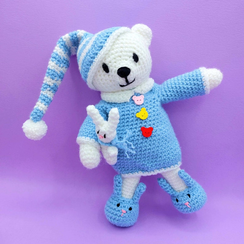 May include: A white crocheted bear wearing a blue and white striped nightcap with a pom-pom, a blue sweater with three bear-shaped buttons in pink, yellow, and red, and blue bunny-shaped slippers. The bear holds a small white crocheted bunny toy.