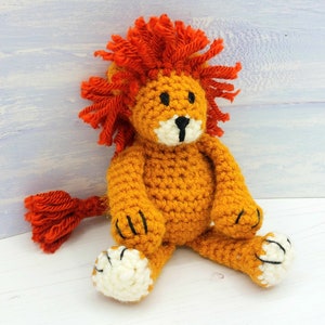 Puede incluir: Un juguete de le&oacute;n de crochet con una melena naranja brillante, un cuerpo amarillo y patas blancas. El le&oacute;n est&aacute; sentado sobre una superficie blanca.