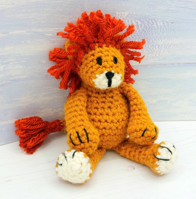 Puede incluir: Un juguete de le&oacute;n de crochet con una melena naranja brillante, un cuerpo amarillo y patas blancas. El le&oacute;n est&aacute; sentado sobre una superficie blanca.
