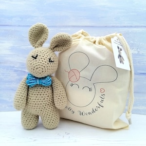 May include: A beige crocheted bunny with blue bow tie, sitting next to a white drawstring bag with a black and red bunny illustration and the text "Olly Wonderfuls".