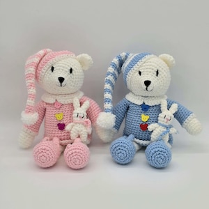 May include: Two crocheted teddy bears, one in pink and white and one in blue and white, each wearing a matching striped nightcap and pajamas with three colorful buttons. Each bear holds a small white crocheted rabbit toy on its chest.